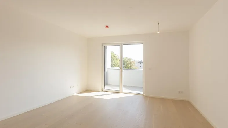 Helles, leeres Zimmer mit Parkettboden und Zugang zu einem sonnigen Balkon.
