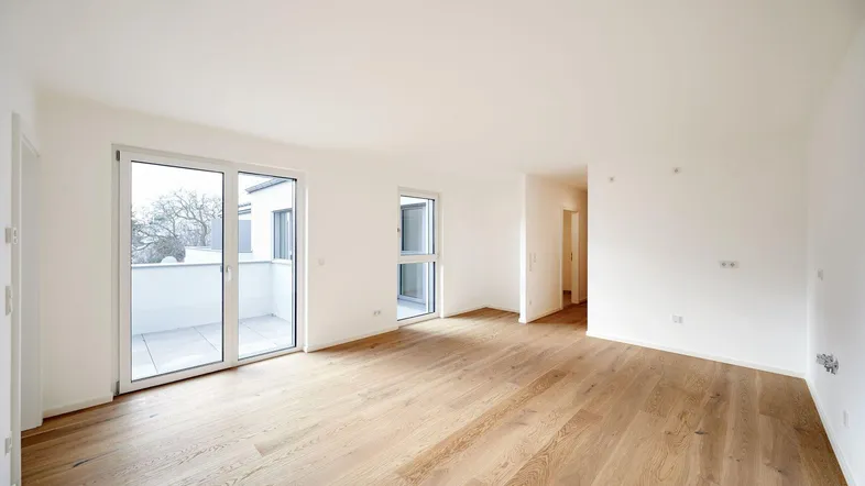 Helles, leeres Wohnzimmer mit modernem Parkettboden und Zugang zu einem Balkon.