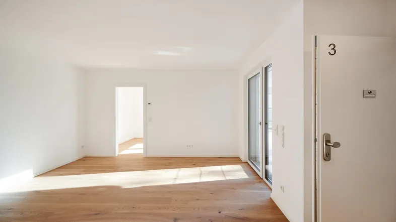 Helles, leeres Zimmer mit Holzboden und Zugang zu einem Außenbereich durch eine Schiebetür.