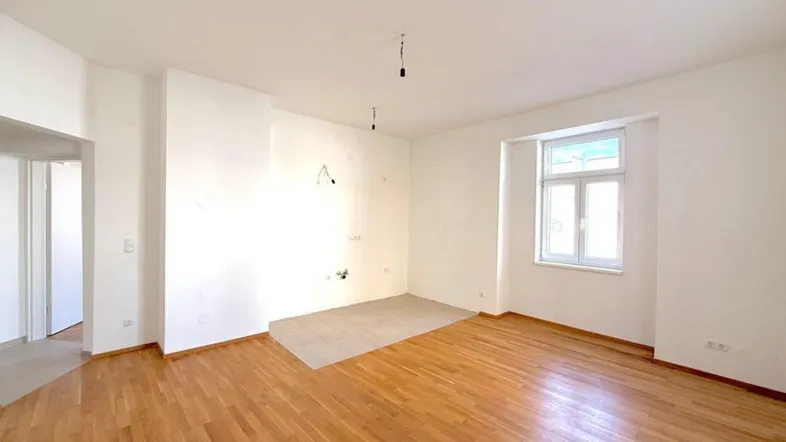 Helles, leeres Zimmer mit Holzboden und vorbereitetem Bereich für eine Küche.