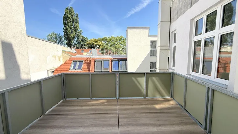Heller Balkon mit modernem Holzboden und Blick auf umliegende Gebäude und Bäume unter blauem Himmel.