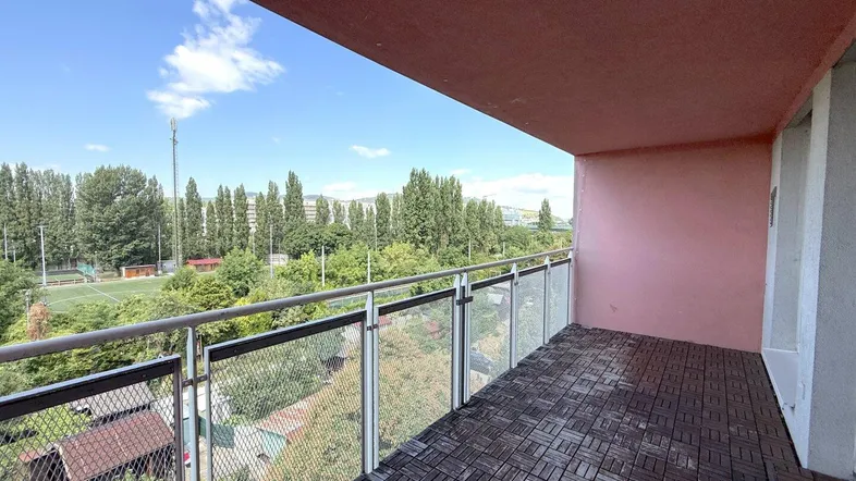 Geräumiger Balkon mit Holzbodenfliesen und weitem Blick ins Grüne sowie auf einen Sportplatz.