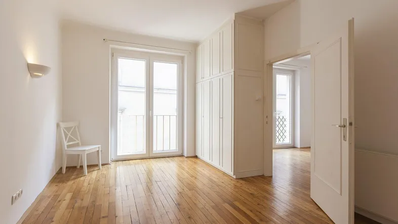Helles Zimmer mit Holzdielenboden, Einbauschrank und Zugang zu einem französischen Balkon.