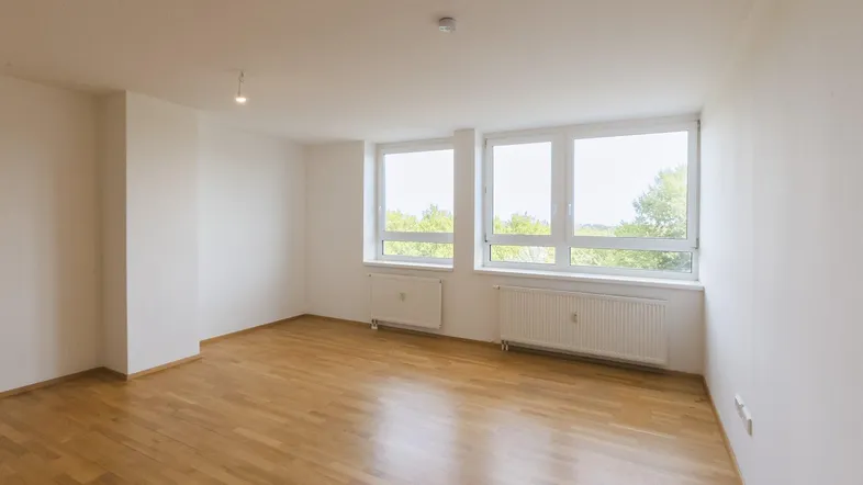 Helles, leeres Zimmer mit Parkettboden und großen Fenstern, die viel Tageslicht hereinlassen.