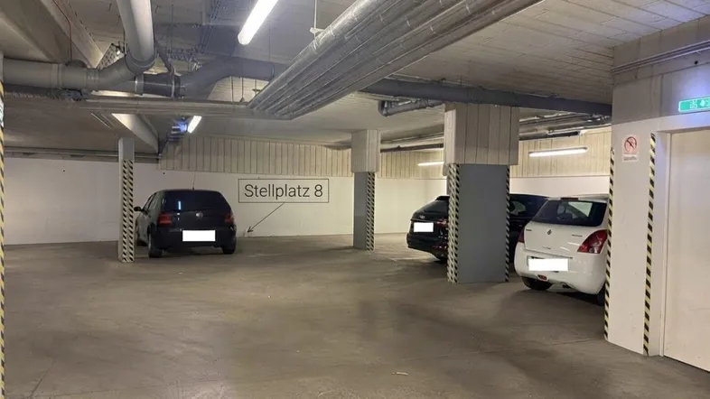 Tiefgarage mit mehreren Stellplätzen, Betonboden, sichtbaren Rohren und Neonbeleuchtung an der Decke.