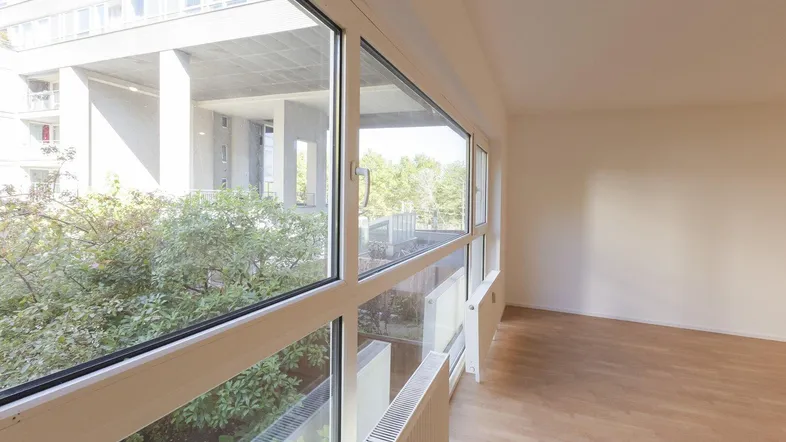 Heller, leerer Raum mit großen Fenstern und Blick ins Grüne, ideal als Wohnzimmer nutzbar.
