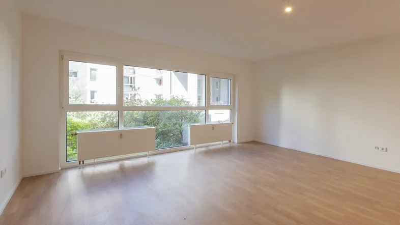Helles, leeres Wohnzimmer mit großen Fenstern, Laminatboden und Blick ins Grüne.