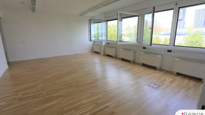 Leeres Büro mit hellem Holzboden und großen, geöffneten Fenstern, die viel Tageslicht und frische Luft hereinlassen.