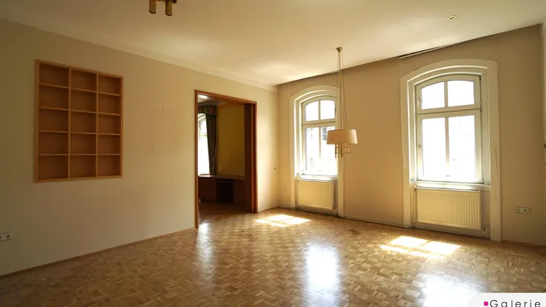 Helles Wohnzimmer mit Fischgrätparkett, hohen Fenstern und einem Durchgang zu einem weiteren Raum.