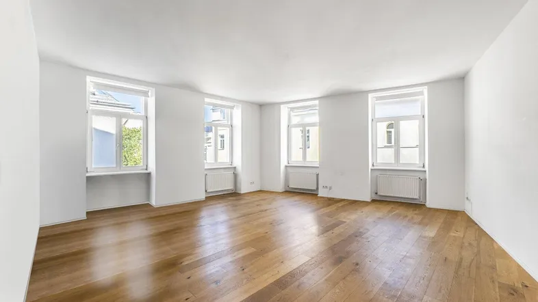 Leeres Wohnzimmer mit glänzendem Holzparkett und mehreren Fenstern, die viel Licht hereinlassen.
