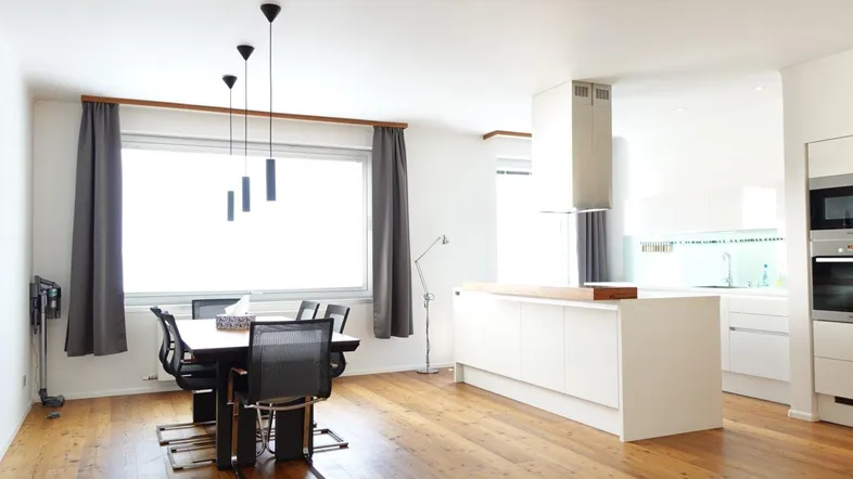 Moderne offene Küche mit weißer Kücheninsel, Essbereich und Holzboden unter großen Fenstern.