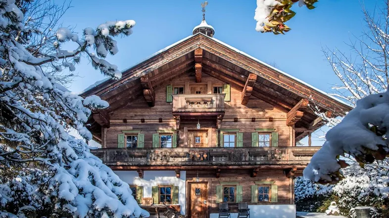 Traditionelles Holzchalet mit verschneitem Garten und Balkonen unter blauem Winterhimmel.