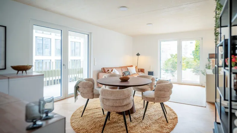 Helles Wohn- und Esszimmer mit Holzboden, runder Teppich und Zugang zu einem Balkon.