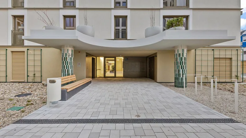 Moderner Eingangsbereich eines Wohngebäudes mit gepflastertem Weg, Sitzbank und dekorativen Säulen.