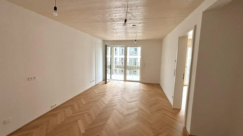 Helles, leeres Zimmer mit Fischgrätparkett und Zugang zu einem Balkon, ideal als Wohnbereich.