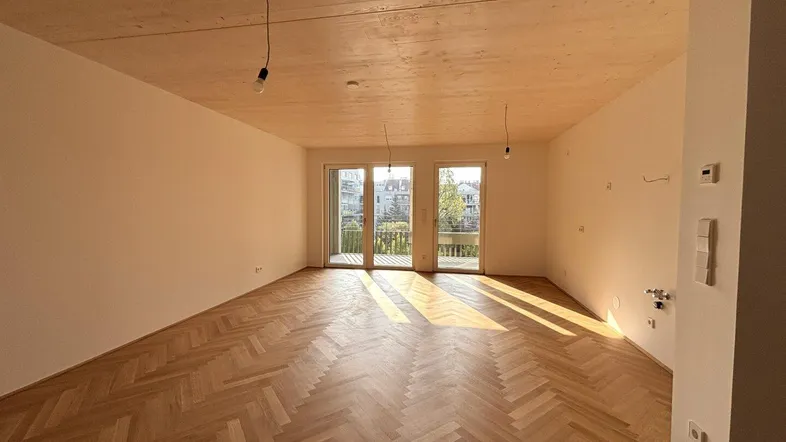 Helles Zimmer mit Fischgrätparkett, Holzdecke und Zugang zu einem Balkon, durchflutet von Sonnenlicht.