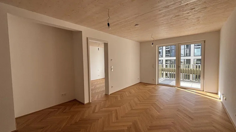 Heller, unmöblierter Wohnraum mit Fischgrätparkettboden, Holzdecke und Zugang zum Balkon.
