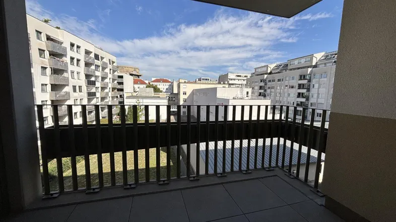 Geräumiger Balkon mit Fliesenboden und Blick auf umliegende Wohngebäude und Grünflächen unter blauem Himmel.