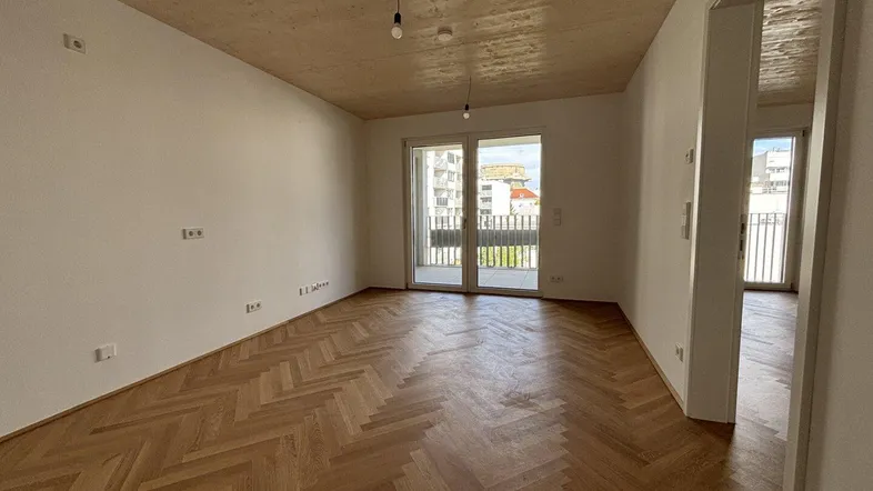 Heller, leerer Wohnraum mit Fischgrätparkett, Holzdecke und Zugang zu einem Balkon in einem Neubau.
