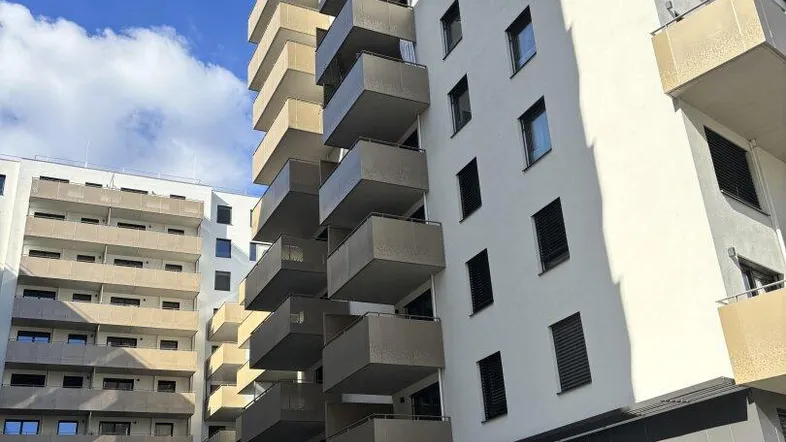 Modernes Mehrfamilienhaus mit zahlreichen Balkonen und gepflegter Grünanlage unter blauem Himmel.