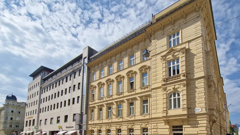 Historisches Gebäude mit gelber Fassade und modernen Anbauten in einer städtischen Umgebung.