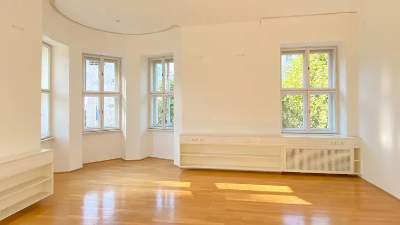 Heller Büroraum mit Holzboden, großen Fenstern und eingebauten Regalen unter den Fenstern.