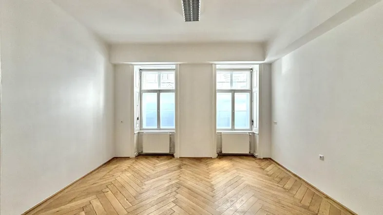 Helles, leeres Zimmer mit klassischem Fischgrätparkett und zwei hohen Fenstern, bereit zur individuellen Gestaltung.
