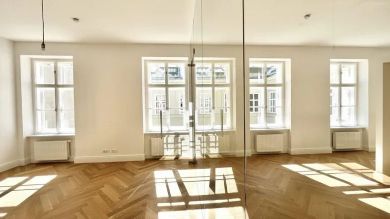 Lichtdurchfluteter Raum mit elegantem Fischgrätparkett und großen Fenstern, die viel Tageslicht hereinlassen.