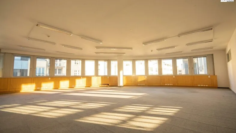 Großer, heller Büroraum mit vielen Fenstern, Teppichboden und integrierten Holzverkleidungen entlang der Wand.