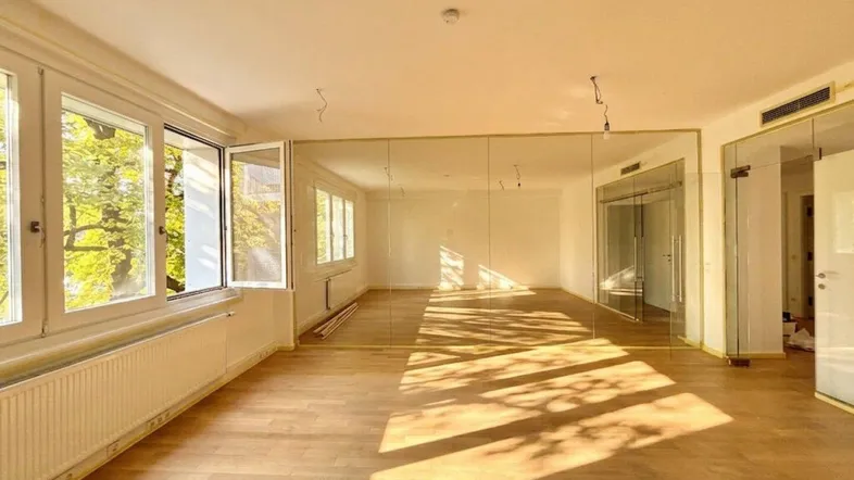 Geräumiger, unmöblierter Wohnbereich mit großen Fenstern, Holzfußboden und einer verspiegelten Wand.