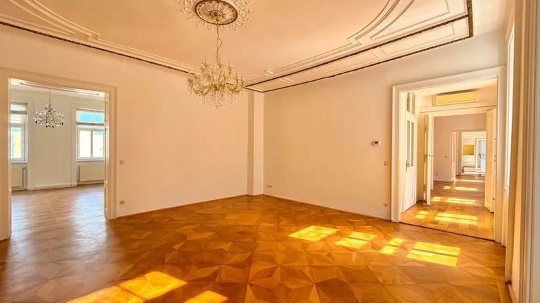 Geräumiges Wohnzimmer mit Fischgrätparkett, hohen Decken und einem eleganten Kronleuchter, durchflutet von natürlichem Licht.