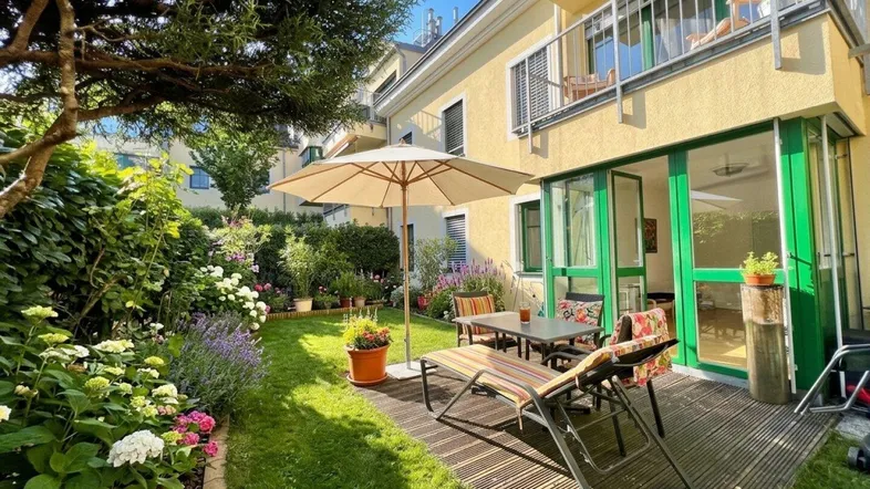 Sonniger Gartenbereich mit Holzterrasse, Gartenmöbeln und üppiger Bepflanzung neben dem Gebäude.