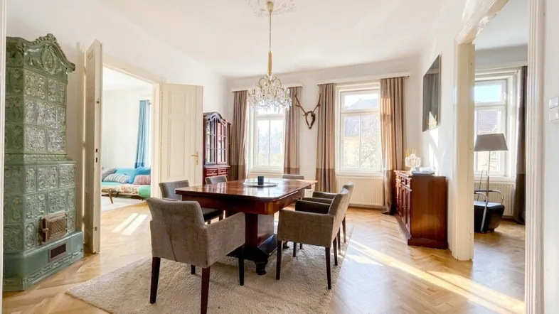 Geräumiges Esszimmer mit elegantem Holztisch, Kachelofen und Fischgrätparkett, durchflutet von Sonnenlicht.
