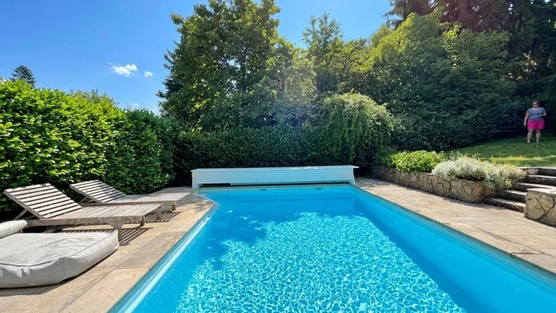 Erfrischender Swimmingpool mit bequemen Liegen, umgeben von grünen Hecken und Bäumen an einem sonnigen Tag.