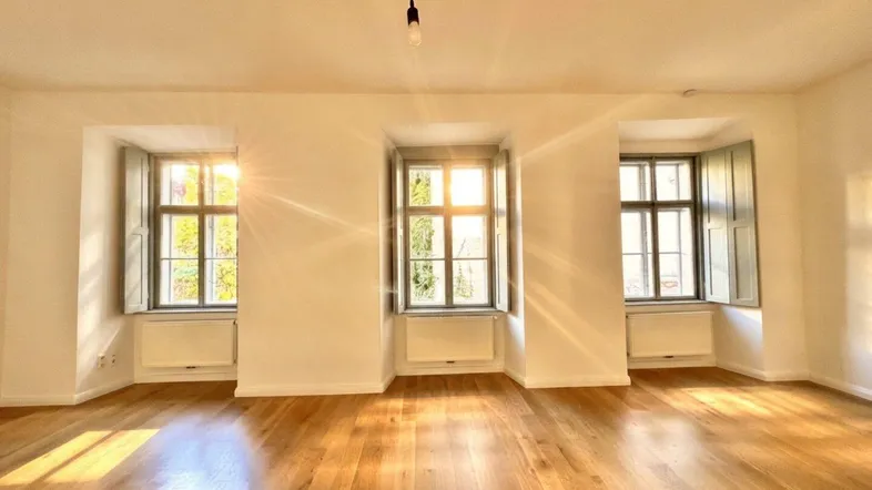 Helles Zimmer mit Holzboden und drei Fenstern, die viel Sonnenlicht hereinlassen.