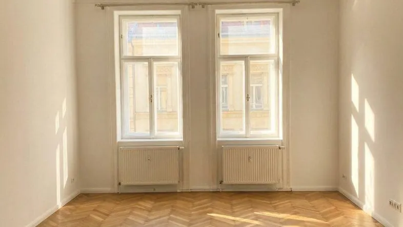 Geräumiges Wohnzimmer mit Fischgrätparkett, hohen Decken und zwei großen Fenstern, die viel Tageslicht hereinlassen.