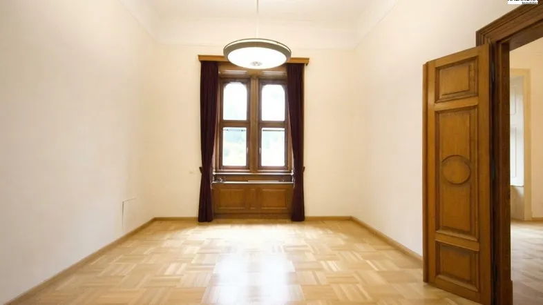 Geräumiger Raum mit hellem Parkettboden, hohen Decken und einem großen Fenster mit dunklen Vorhängen.