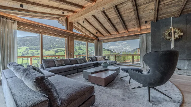 Elegantes Wohnzimmer mit großzügiger Couch, Holzbalkendecke und weitem Blick auf die Alpenlandschaft.