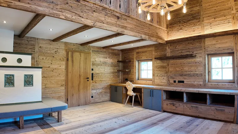 Geräumiges Wohnzimmer im alpinen Stil mit Holzbalken, Kachelofen und einem Arbeitsbereich.