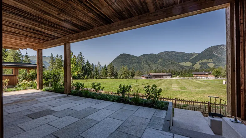 Überdachte Terrasse mit Steinplattenboden und weitem Blick auf grüne Felder und Berge.
