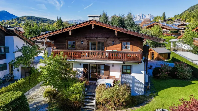 Traditionelles Alpenhaus mit Holzbalkonen, gepflegtem Garten und Bergblick unter blauem Himmel.