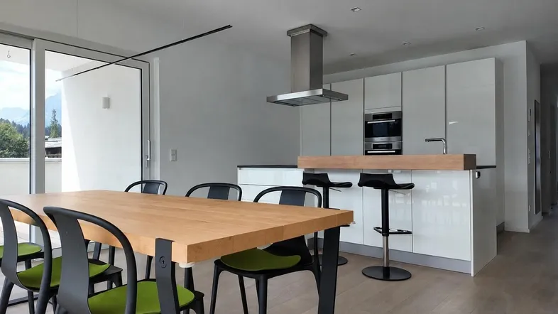 Moderner offener Wohn-Essbereich mit weißer Küche, Holztisch und Zugang zum Balkon mit Bergblick.