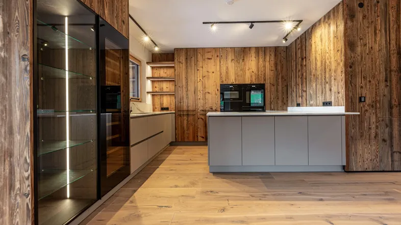 Moderne Küche mit Holzwänden, hellgrauen Schränken, Kochinsel und integrierten Geräten, ideal für alpines Wohnen.