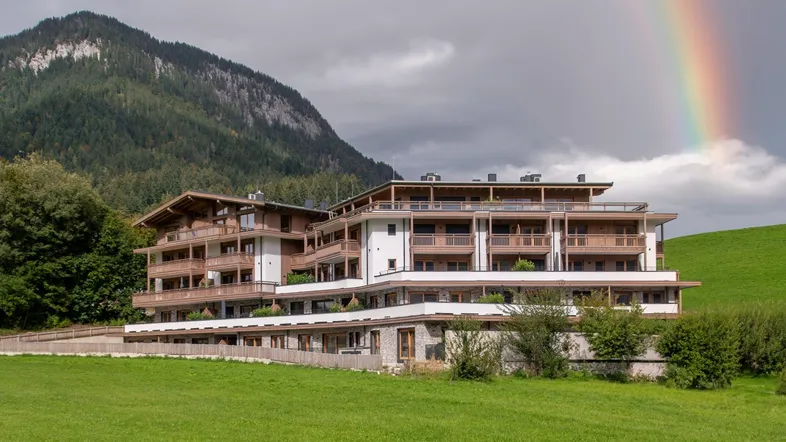 Modernes Gebäude im alpinen Stil mit Balkonen, umgeben von grünen Wiesen und Bergen, mit einem Regenbogen am Himmel.