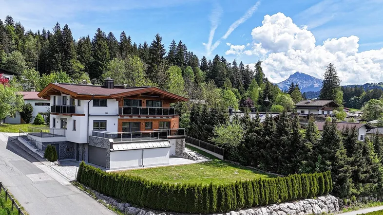 Modernes Haus mit Holzfassade, Balkonen und gepflegtem Garten vor alpiner Bergkulisse.