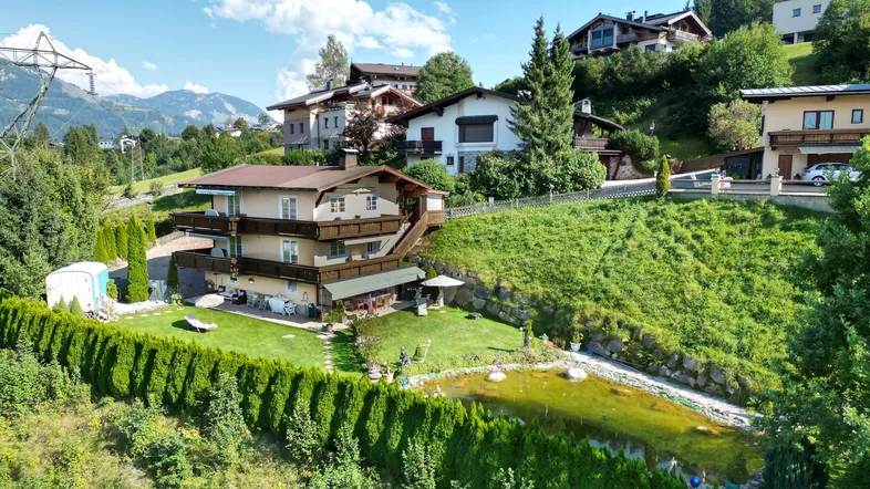 Luftaufnahme eines mehrstöckigen Hauses am Hang mit Garten, Teich und Bergblick.