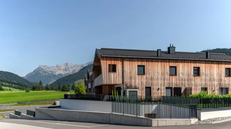 Modernes Holzhaus mit Bergblick und Zufahrt, umgeben von grünen Wiesen unter blauem Himmel.