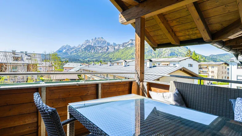 Sonniger Balkon mit Holzbalken, Outdoor-Möbeln und herrlichem Bergblick über die Dächer des Dorfes.