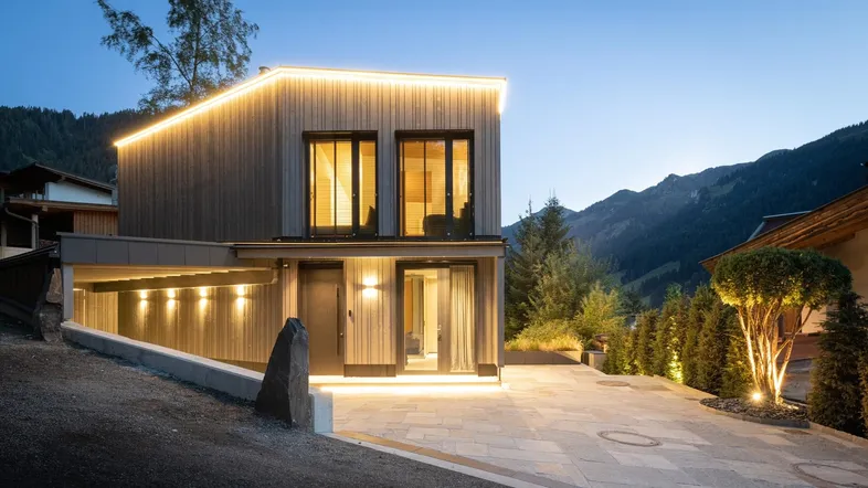Modernes Haus bei Dämmerung mit beleuchteter Holzfassade und Bergblick in alpiner Umgebung.