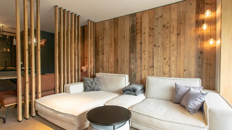 Gemütliches Wohnzimmer mit bequemer Couch, Holzwandverkleidung und dekorativen Holzlamellen als Raumteiler.
